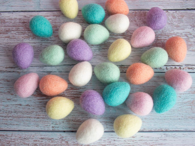 May include: A collection of colourful felt Easter eggs in various pastel shades, including pink, blue, green, yellow, and white. The eggs are arranged on a light wood background.