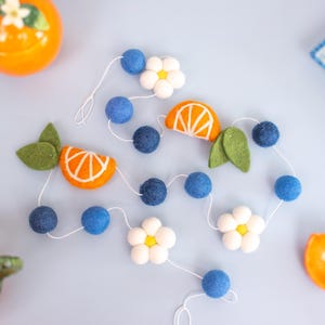 May include: A felt garland featuring dark blue spheres, white five-petaled flowers with yellow centers, orange citrus slices with white detailing, and green leaves strung together on a thin white cord, arranged in a loose, winding pattern.
