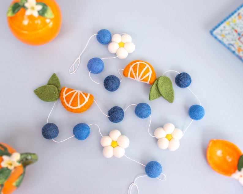 May include: A felt garland featuring dark blue spheres, white five-petaled flowers with yellow centers, orange citrus slices with white detailing, and green leaves strung together on a thin white cord, arranged in a loose, winding pattern.