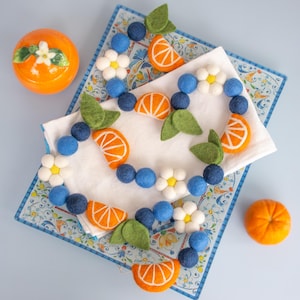 May include: A felt ball garland featuring blue spheres, white flowers with yellow centers, orange citrus slice shapes, and green leaves. The garland is arranged on a white cloth atop a rectangular tray with a blue and white floral pattern.