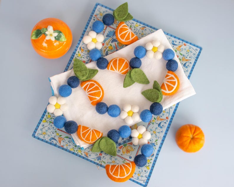 May include: A felt ball garland featuring blue spheres, white flowers with yellow centers, orange citrus slice shapes, and green leaves. The garland is arranged on a white cloth atop a rectangular tray with a blue and white floral pattern.