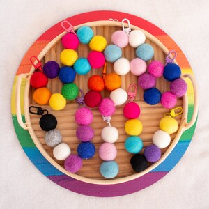 May include: A rainbow assortment of felt balls with metal rings attached, displayed on a wooden tray with a rainbow-colored rim. The felt balls are arranged in a circle, with the metal rings facing outwards.