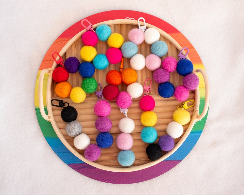 May include: A rainbow assortment of felt balls with metal rings attached, displayed on a wooden tray with a rainbow-colored rim. The felt balls are arranged in a circle, with the metal rings facing outwards.