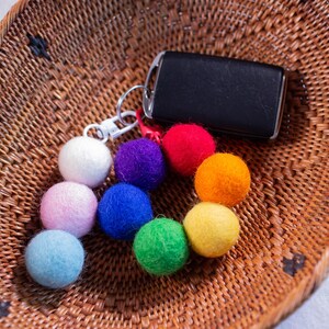 May include: A keychain with a black car key and a colorful set of felt balls in rainbow colors, including red, orange, yellow, green, blue, and purple. The keychain is sitting on a woven basket.