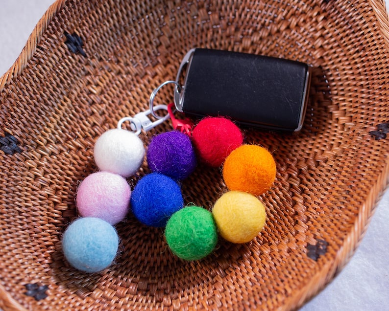 May include: A keychain with a black car key and a colorful set of felt balls in rainbow colors, including red, orange, yellow, green, blue, and purple. The keychain is sitting on a woven basket.