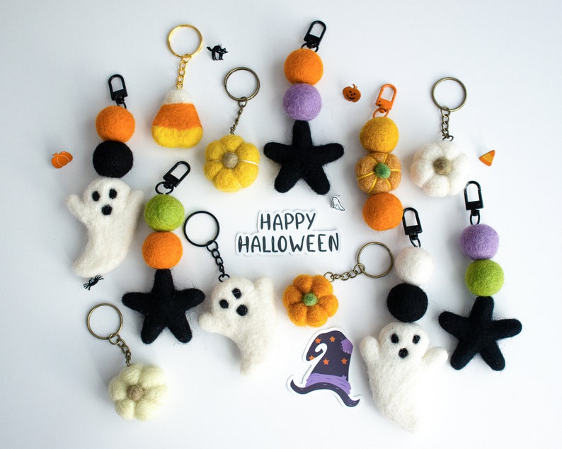 K&ouml;nnte beinhalten: Eine Sammlung von Halloween-Schl&uuml;sselanh&auml;ngern aus Filz mit Geistern, Sternen, K&uuml;rbissen, Zuckermais und Blumen. Die Schl&uuml;sselanh&auml;nger sind an goldenen Ketten befestigt und haben schwarze und wei&szlig;e Filzaugen. Der Text "Happy Halloween" ist auf einem wei&szlig;en Aufkleber geschrieben.