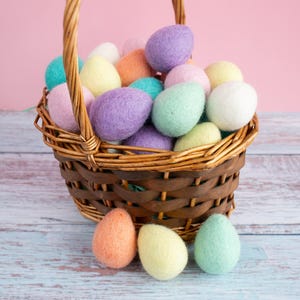 May include: A wicker basket filled with colourful felt Easter eggs. The basket is sitting on a light blue wooden surface. There are four felt eggs outside of the basket.