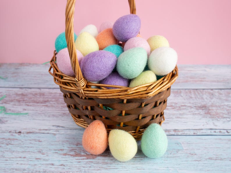 May include: A wicker basket filled with colourful felt Easter eggs. The basket is sitting on a light blue wooden surface. There are four felt eggs outside of the basket.