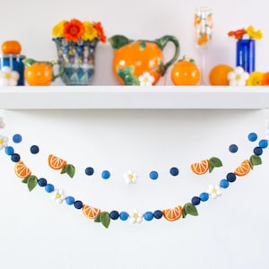 May include: A felt ball garland featuring orange slices, white flowers, and dark blue spheres. The garland is draped in two rows, showcasing a repeating pattern of citrus segments, green leaves, and small white blossoms strung together.