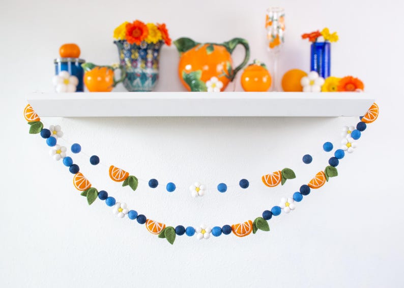 May include: A felt ball garland featuring orange slices, white flowers, and dark blue spheres. The garland is draped in two rows, showcasing a repeating pattern of citrus segments, green leaves, and small white blossoms strung together.