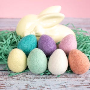 May include: Seven felt Easter eggs in various pastel colours, including yellow, green, blue, purple, pink, and orange, are arranged on a light blue wooden surface. The eggs are surrounded by green shredded paper and a white ceramic bunny.