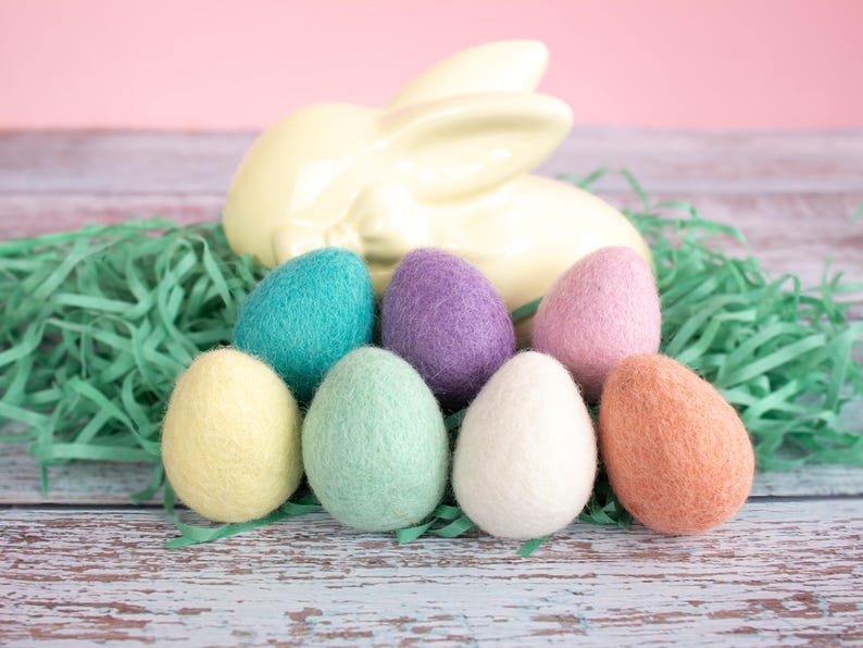 May include: Seven felt Easter eggs in various pastel colours, including yellow, green, blue, purple, pink, and orange, are arranged on a light blue wooden surface. The eggs are surrounded by green shredded paper and a white ceramic bunny.