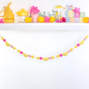 May include: A yellow and pink felt garland with lemon slices and white pom poms. The garland is hanging on a white shelf with a variety of yellow and pink decor.