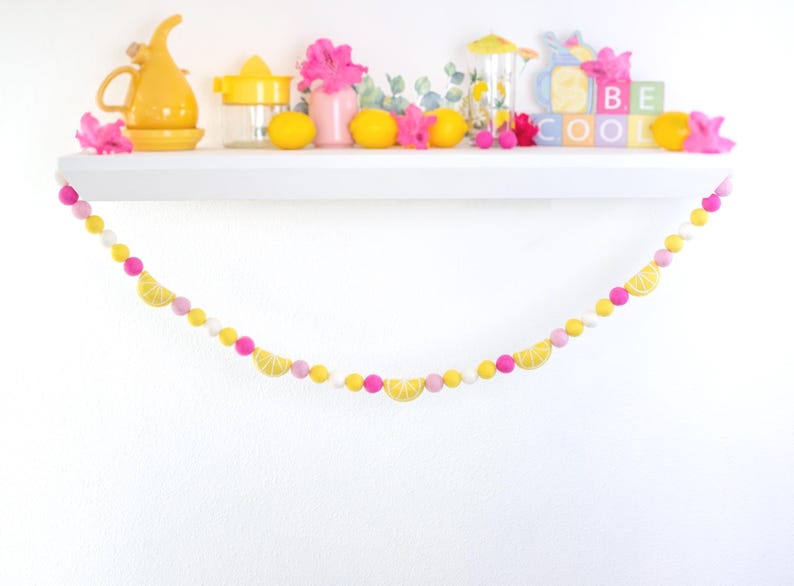 May include: A yellow and pink felt garland with lemon slices and white pom poms. The garland is hanging on a white shelf with a variety of yellow and pink decor.