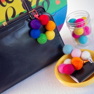 May include: A black leather handbag with a rainbow pom-pom keychain. The keychain is attached to the bag with a gold metal clasp. The bag is sitting on a white surface.