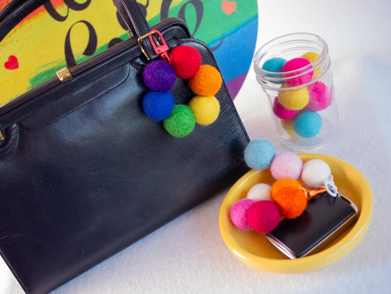 May include: A black leather handbag with a rainbow pom-pom keychain. The keychain is attached to the bag with a gold metal clasp. The bag is sitting on a white surface.