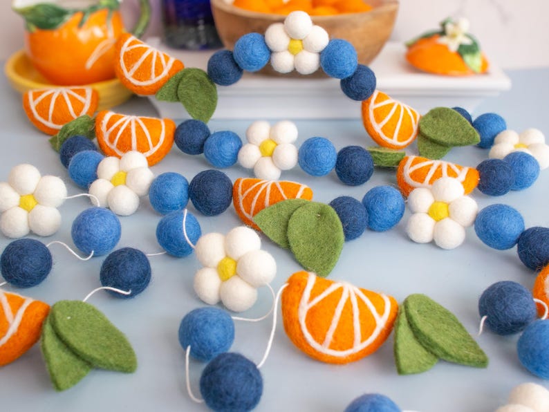 May include: A felt garland featuring orange citrus slices, white flowers with yellow centers, dark blue and light blue spheres, and green leaves. The felt elements are strung together on a white cord, creating a repeating pattern of fruit, floral, and spherical shapes.