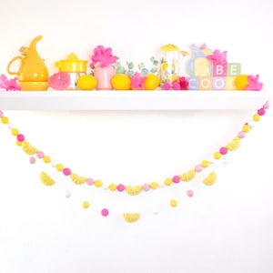 May include: A yellow and pink felt ball garland with lemon slices hanging from it. The garland is hanging from a white shelf with yellow lemons, pink flowers, and a yellow pitcher on it. The shelf also has a sign that says "BE COOL".