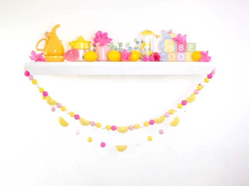 May include: A yellow and pink felt ball garland with lemon slices hanging from it. The garland is hanging from a white shelf with yellow lemons, pink flowers, and a yellow pitcher on it. The shelf also has a sign that says "BE COOL".