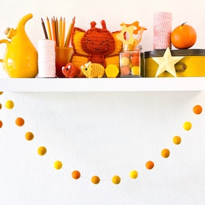 May include: A white shelf with a yellow ceramic pitcher, a spool of white yarn, a jar of colored pencils, a crocheted butterfly, a crocheted bee, a jar of yellow beads, a yellow wooden box, a yellow star, and a garland of yellow and orange felt balls hanging from the shelf.