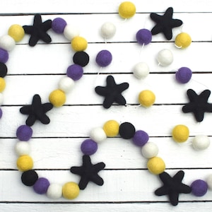 May include: A decorative garland featuring black star shapes and colorful felt balls in yellow, purple, white, and black. The garland is strung on a white string and is displayed on a white wooden surface.