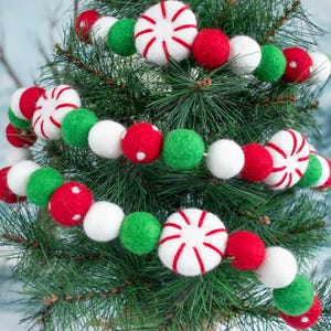 May include: A festive Christmas garland with red, white, and green felt balls and peppermint candy ornaments. The garland is wrapped around a green Christmas tree.