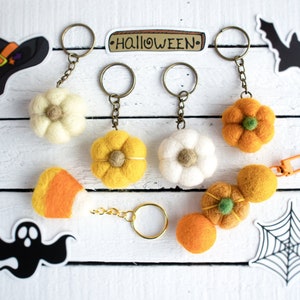 K&ouml;nnte beinhalten: F&uuml;nf Filz-Halloween-Schl&uuml;sselanh&auml;nger mit verschiedenen Designs. Die Schl&uuml;sselanh&auml;nger beinhalten einen wei&szlig;en K&uuml;rbis, einen gelben K&uuml;rbis, ein Zuckermais, ein wei&szlig;es Gespenst und ein Spinnennetz. Die Schl&uuml;sselanh&auml;nger sind an goldenen Schl&uuml;sselringen befestigt. Die Schl&uuml;sselanh&auml;nger befinden sich auf einer wei&szlig;en Holzoberfl&auml;che mit einem Schild, auf dem "HALLOWEEN" steht.