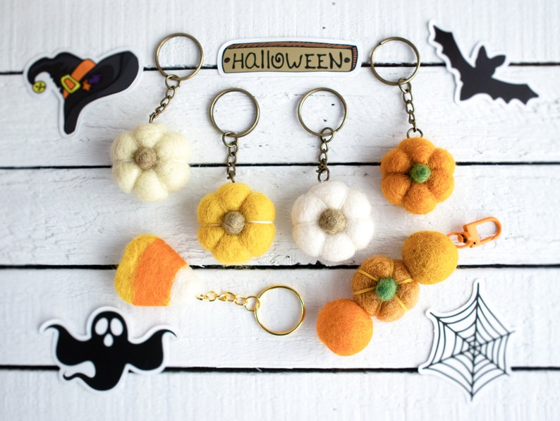K&ouml;nnte beinhalten: F&uuml;nf Filz-Halloween-Schl&uuml;sselanh&auml;nger mit verschiedenen Designs. Die Schl&uuml;sselanh&auml;nger beinhalten einen wei&szlig;en K&uuml;rbis, einen gelben K&uuml;rbis, ein Zuckermais, ein wei&szlig;es Gespenst und ein Spinnennetz. Die Schl&uuml;sselanh&auml;nger sind an goldenen Schl&uuml;sselringen befestigt. Die Schl&uuml;sselanh&auml;nger befinden sich auf einer wei&szlig;en Holzoberfl&auml;che mit einem Schild, auf dem "HALLOWEEN" steht.