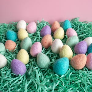 May include: A collection of pastel coloured felt Easter eggs in a bed of green shredded paper. The eggs are in various shades of pink, blue, green, and yellow.