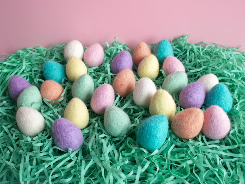 May include: A collection of pastel coloured felt Easter eggs in a bed of green shredded paper. The eggs are in various shades of pink, blue, green, and yellow.