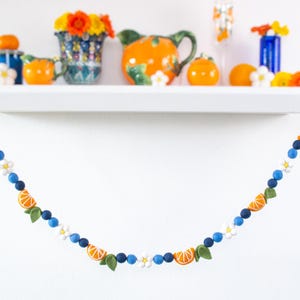 May include: A decorative garland featuring felt orange slices, small green leaves, white flowers, and alternating dark and light blue felt balls. The garland is draped in a curved shape, showcasing a repeating pattern of citrus and floral elements.