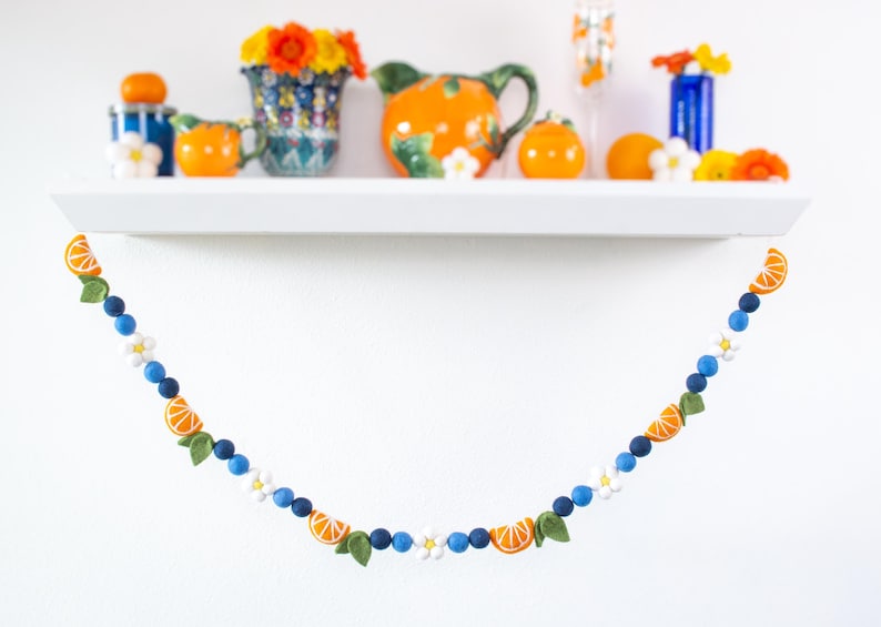 May include: A decorative garland featuring felt orange slices, small green leaves, white flowers, and alternating dark and light blue felt balls. The garland is draped in a curved shape, showcasing a repeating pattern of citrus and floral elements.