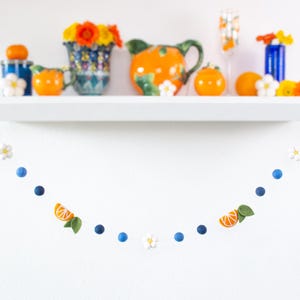 May include: A decorative garland featuring felted blue spheres, white five-petaled flowers, and orange citrus slice segments with green leaves, strung together in a repeating pattern and suspended in a curved shape against a white wall.