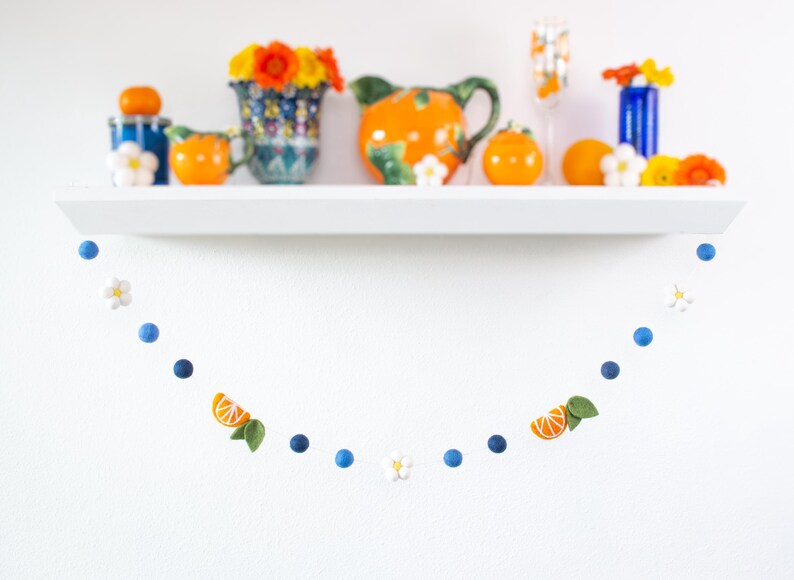 May include: A decorative garland featuring felted blue spheres, white five-petaled flowers, and orange citrus slice segments with green leaves, strung together in a repeating pattern and suspended in a curved shape against a white wall.