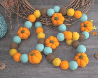 Op de afbeelding: Een herfstthema slinger met vilten pompoenen en kleurrijke ballen in oranje, geel, bruin en blauwgroen. De slinger is in een decoratief patroon gerangschikt, perfect voor herfstdecoratie. Achtergrond met een gevlochten krans.