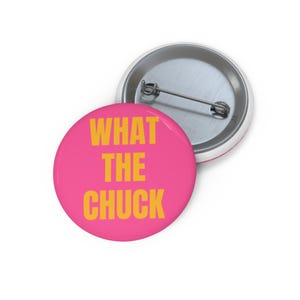 Op de afbeelding: Een ronde button met een felroze achtergrond en de tekst WHAT THE CHUCK in dikgedrukte, gele hoofdletters. De achterkant van de button is van zilverkleurig metaal en is voorzien van een standaard speldmechanisme voor bevestiging.