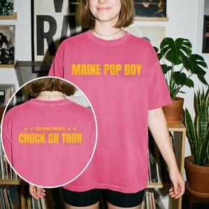 Op de afbeelding: Een roze t-shirt met korte mouwen en de gele tekst MAINE POP BOY op de voorkant. Een ingevoegde cirkel toont de achterkant van hetzelfde t-shirt met de gele tekst ROLE MODEL PRESENTS CHUCK ON TOUR en sterpictogrammen boven de tekst.