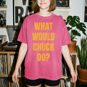 May include: A short-sleeve crew neck t-shirt in a faded pink color. The front of the shirt features the text WHAT WOULD CHUCK DO? printed in large, bold, yellow capital letters centered on the chest. The garment has a relaxed, loose fit.