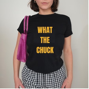 May include: A black short-sleeved t-shirt featuring the yellow text WHAT THE CHUCK printed on the front. The shirt is paired with a bright pink handbag with a textured crocodile-skin pattern and black and white gingham patterned bottoms.
