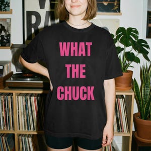 Op de afbeelding: Een zwart T-shirt met korte mouwen en de tekst WHAT THE CHUCK in vetgedrukte, felroze hoofdletters op de borst. Het kledingstuk heeft een klassieke ronde hals en een relaxte pasvorm, wat zorgt voor een eenvoudig en direct grafisch ontwerp.