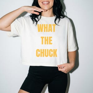 Op de afbeelding: Een crèmekleurig cropped T-shirt met de tekst WHAT THE CHUCK in dikke, gele hoofdletters op de voorzijde. Het kledingstuk heeft korte mouwen en een boxy pasvorm, gecombineerd met een zwarte, nauwsluitende short tot halverwege de dij.