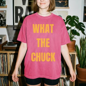 May include: A short-sleeved, crew-neck t-shirt in a faded pink color. The front of the shirt features the text "WHAT THE CHUCK" printed in large, bold, yellow capital letters centered on the chest. The garment has a relaxed, loose-fitting cut.