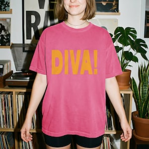 May include: A short-sleeved, crew-neck t-shirt in a faded pink color. The front of the shirt features the word DIVA! printed in large, bold, mustard-yellow capital letters across the chest area. The garment has a relaxed, loose-fitting cut.