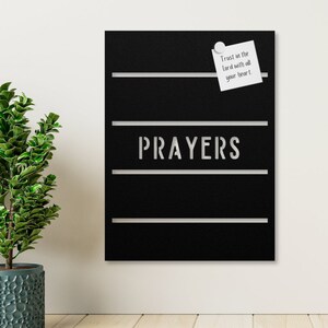 May include: A black metal board with the word "PRAYERS" in silver lettering. The board has four horizontal lines and a white sticky note with the text "Trust in the Lord with all your heart." attached to the top line.