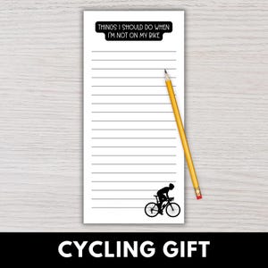 May include: White notepad with horizontal lines, a black silhouette of a cyclist, and the text "THINGS I SHOULD DO WHEN I'M NOT ON MY BIKE." A yellow pencil rests on the notepad. The words "CYCLING GIFT" are at the bottom.