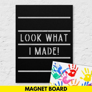 May include: Black magnetic board with white text that reads "Look What I Made!"  The board is mounted on a white wall. A white sheet of paper with colorful handprints is attached to the board with a magnet.