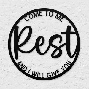 May include: Black metal wall art in a circular design. The words "COME TO ME" are at the top, "Rest" in a cursive font in the center, and "AND I WILL GIVE YOU" at the bottom. The art is mounted on a textured white wall.