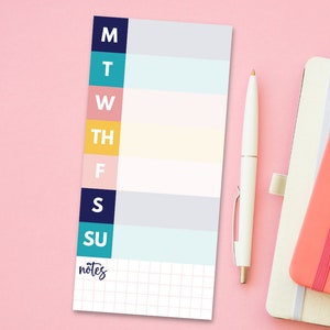 May include: A white weekly planner with colorful tabs for each day of the week. The tabs are labeled M, T, W, TH, F, S, and SU. The planner has a gridded notes section at the bottom.
