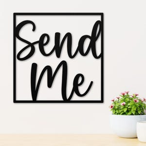 May include: Black wooden wall art with the words "Send Me" in a cursive font, framed by a black square. The art is displayed on a white wall, with a small potted plant in the lower right corner.