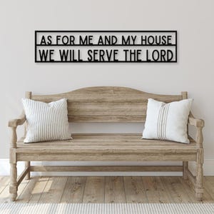 May include: Black metal wall decor with the text "As for me and my house we will serve the Lord". The decor is hanging above a wooden bench with white striped cushions.